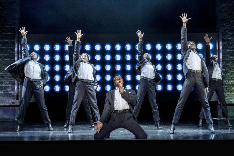 The UK Company of Ain’t Too Proud. Seven men are on stage all wearing matching black and white suits. They are all performing a song with one arm extended up and there are many bright lights behind them. One man is on his knees in the front, singing passionately into a microphone.