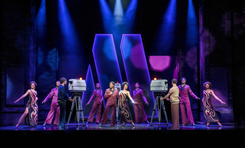 The UK Company of Ain’t Too Proud. Three women stand on stage in long patterned sequin dresses. They are joined by five men wearing matching maroon suits. They perform a song on stage in front of vintage cameras.