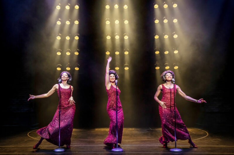 The UK Company of Ain’t Too Proud. Three women are standing side by side wearing long red sequin dresses. They are standing in front of microphone stands performing a song with many bright lights behind them.