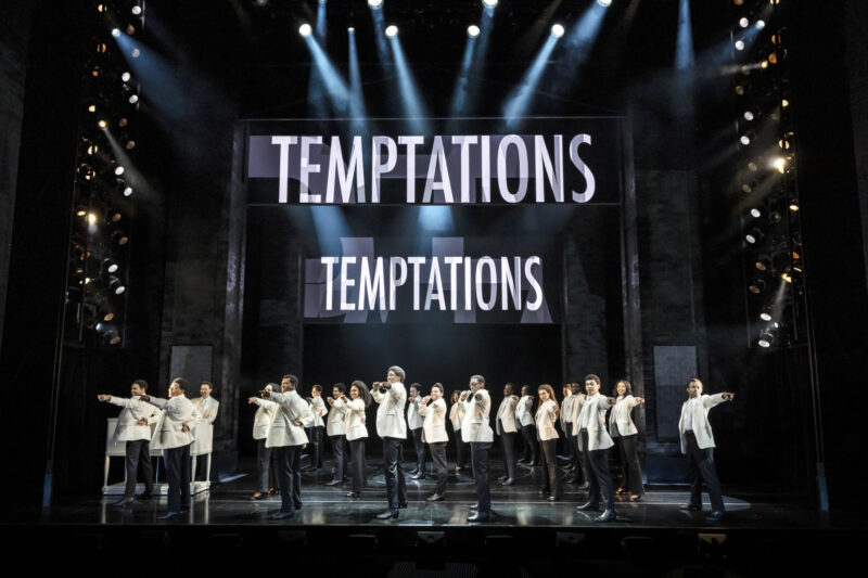 The UK Company of Ain’t Too Proud. A large group of people are all wearing white suit shirts and jackets with black pants They are all turned sideways with their left hands pointed out at the audience. Two signs behind them read “Temptations”.